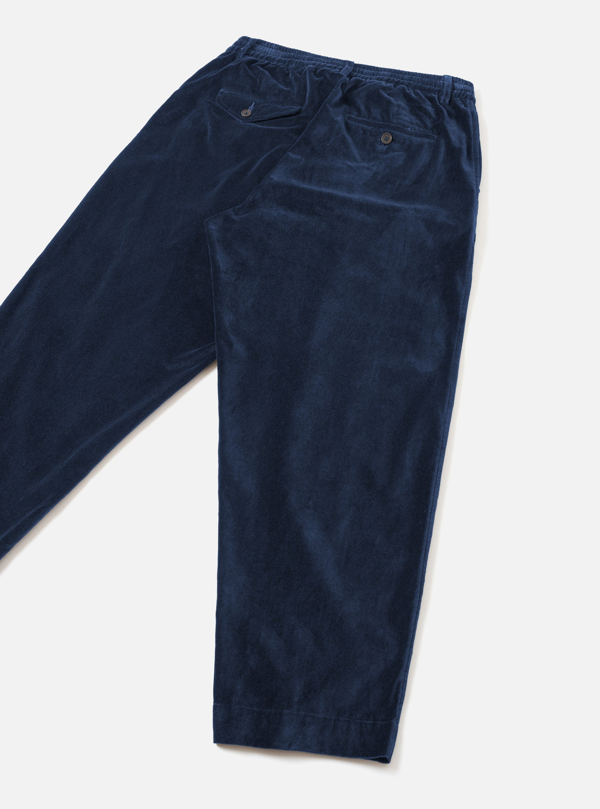 Navy velvet Oxford trousers laid flat on a light grey surface, shot from above at an angle. Fine-wale velvet texture is visible across the fabric. The elasticated waistband is shown at the top, with a buttoned welt pocket on the back and a side pocket visible. The tapered legs end in plain hems.