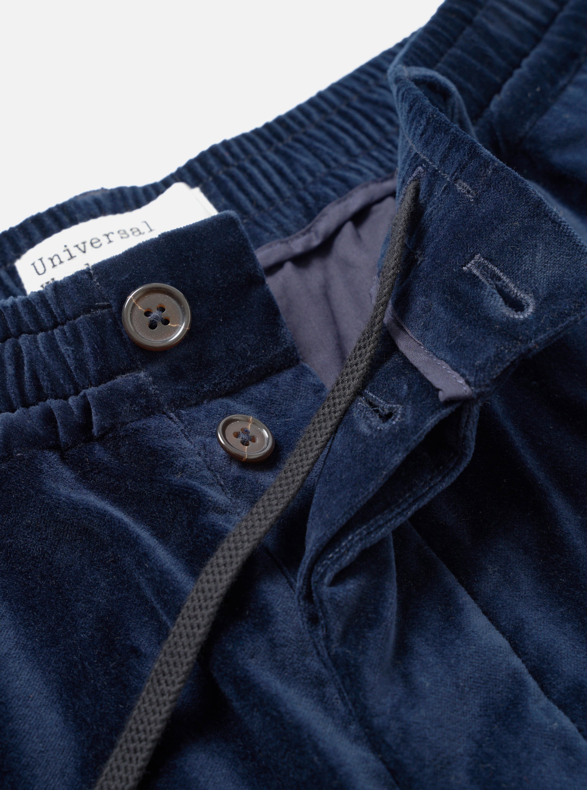 Close-up detail of navy velvet trousers on a plain light grey studio backdrop. The shot focuses on the elasticated waistband, button fly with two dark horn buttons, and a partially open fly revealing a dark grey lining and woven drawcord. A Universal Works label is visible at the waistband interior.