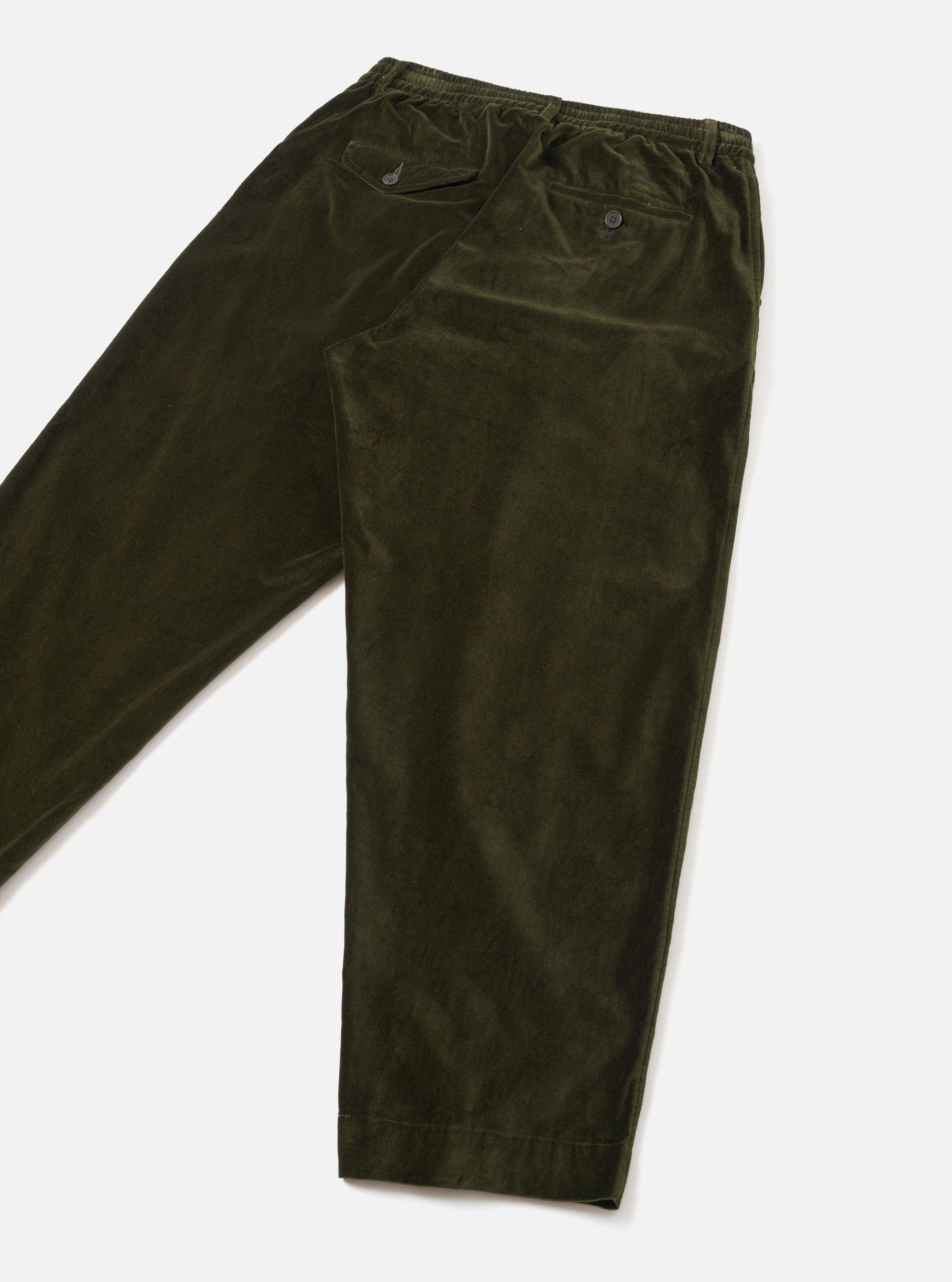 Olive velvet corduroy trousers laid flat on a light grey seamless surface, shot at an angle. The elasticated waistband is visible at the top, with two buttoned flap pockets at the front and a single button-fastening welt pocket at the rear. The legs taper slightly toward plain, unfinished hems.
