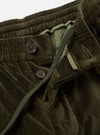 Close-up detail of olive velvet oxford trousers on a white studio backdrop. The elasticated waistband, button fly with two dark brown four-hole buttons, and a woven green drawcord are visible. A pale green woven care label and light olive fabric lining are shown inside the open fly.