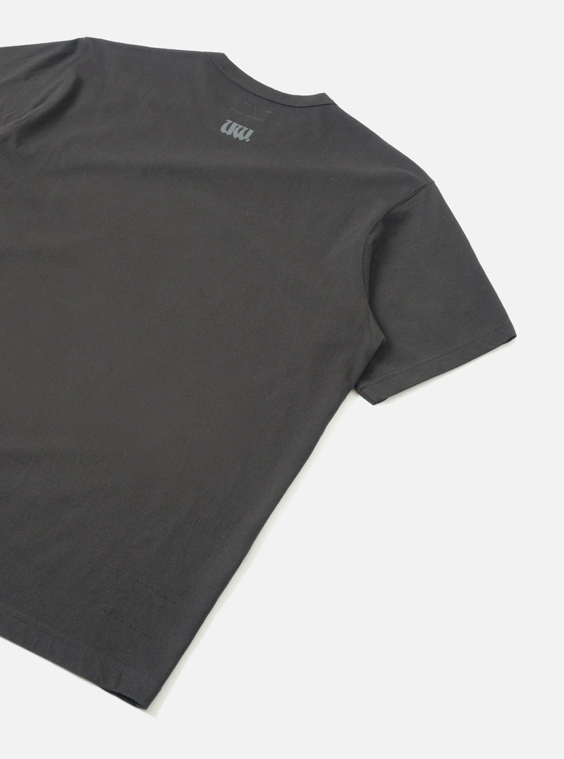 Flat lay of a black single jersey t-shirt shot from behind on a light grey seamless backdrop. The back panel is plain with a small tonal UW logo print near the neckline. Smooth jersey fabric with a clean crew neck and short sleeves with hemmed cuffs visible.