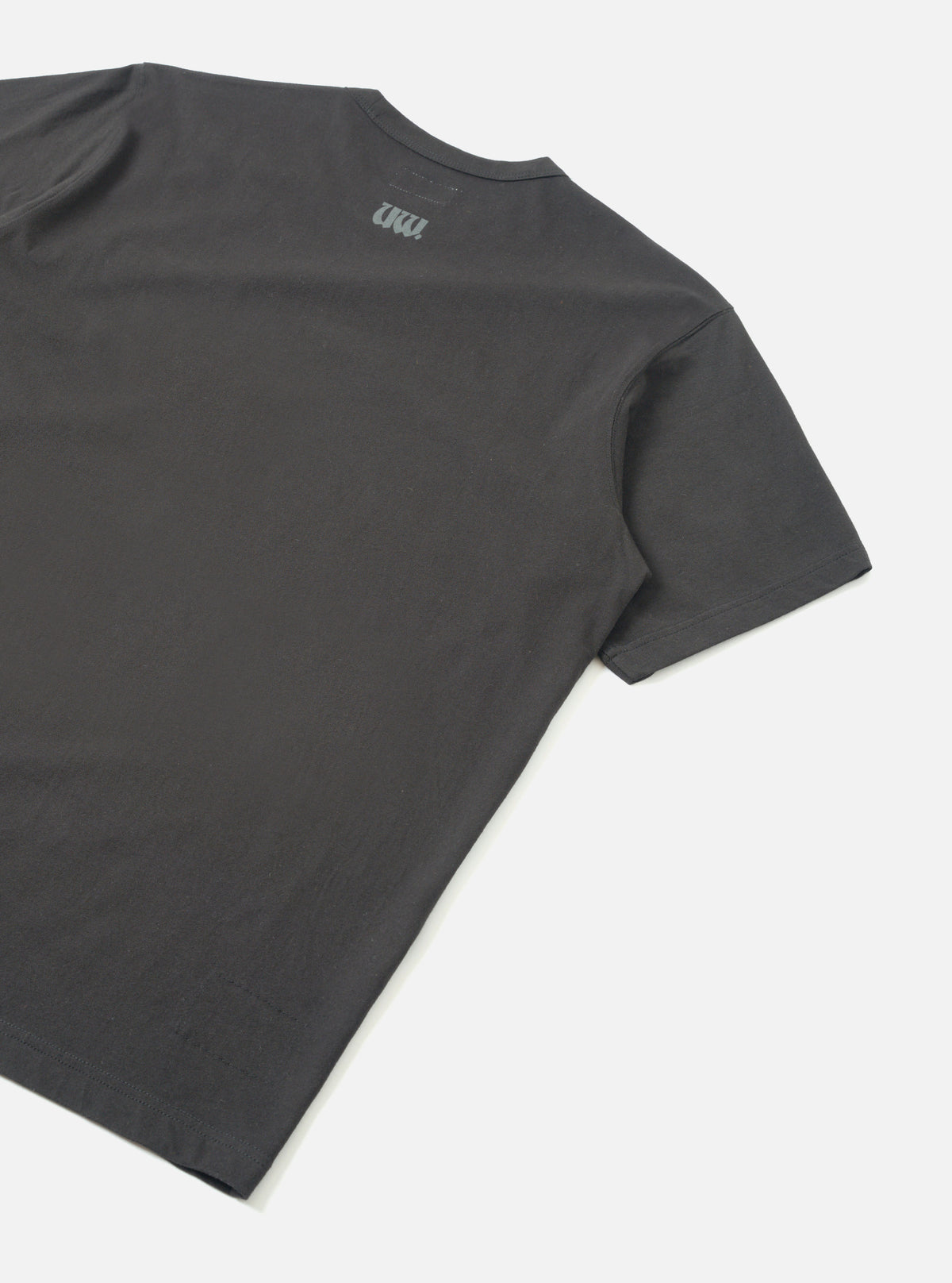 Flat lay of a black single jersey t-shirt shot from behind on a light grey seamless backdrop. The back panel is plain with a small tonal UW logo print near the neckline. Smooth jersey fabric with a clean crew neck and short sleeves with hemmed cuffs visible.