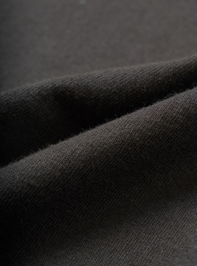 Close-up detail shot of the black single jersey fabric used in the Core Tee, showing a fine knit texture with a soft, slightly raised surface. The material is folded to reveal its weight and drape. Shot against a dark grey seamless backdrop.