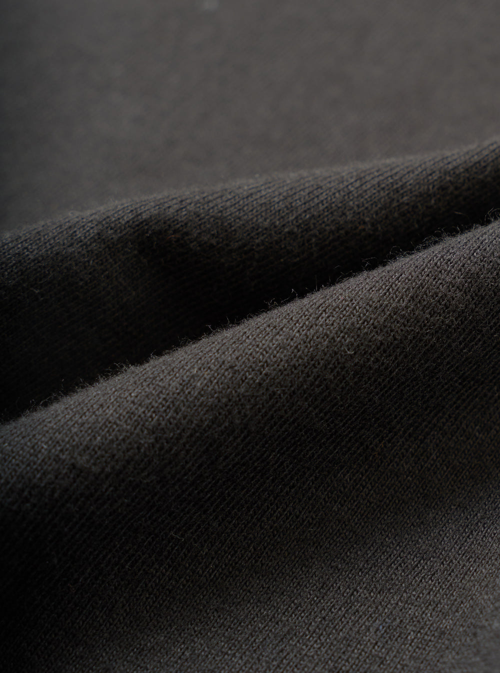 Close-up detail shot of the black single jersey fabric used in the Core Tee, showing a fine knit texture with a soft, slightly raised surface. The material is folded to reveal its weight and drape. Shot against a dark grey seamless backdrop.