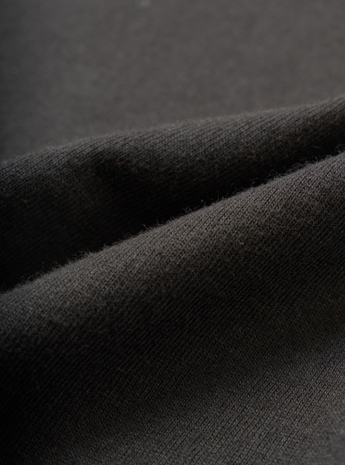 Close-up detail shot of the black single jersey fabric used in the Core Tee, showing a fine knit texture with a soft, slightly raised surface. The material is folded to reveal its weight and drape. Shot against a dark grey seamless backdrop.