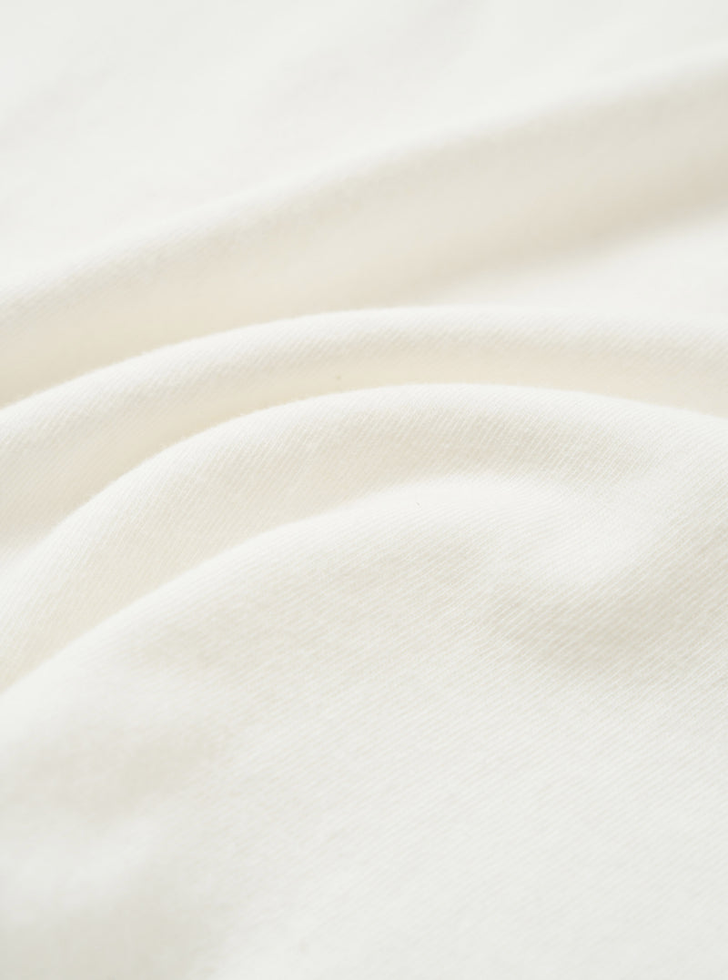 Close-up of the ecru single jersey fabric used in the Core Tee, showing a soft, fine knit texture with natural drape and gentle folds across the frame. The off-white tone is consistent throughout. No garment details or model are visible.