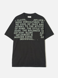 Black short-sleeve t-shirt in single jersey cotton, laid flat on a light grey studio backdrop. Crew neck with a small woven Universal Works label at the back neck. Large sage green bold text print covers the chest, referencing jazz, punk, mods and hip-hop. A plain patch pocket sits at the lower right chest, partially obscuring the print.