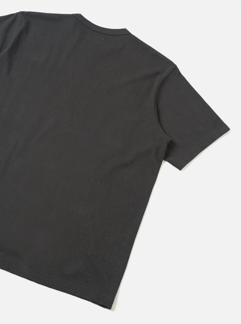 Back view of a black single jersey pocket tee, laid flat on a light grey surface. The shot is cropped to show the upper back, one short sleeve with a plain hem, and a care label visible at the nape of the neck. The fabric is smooth with a fine jersey knit texture.