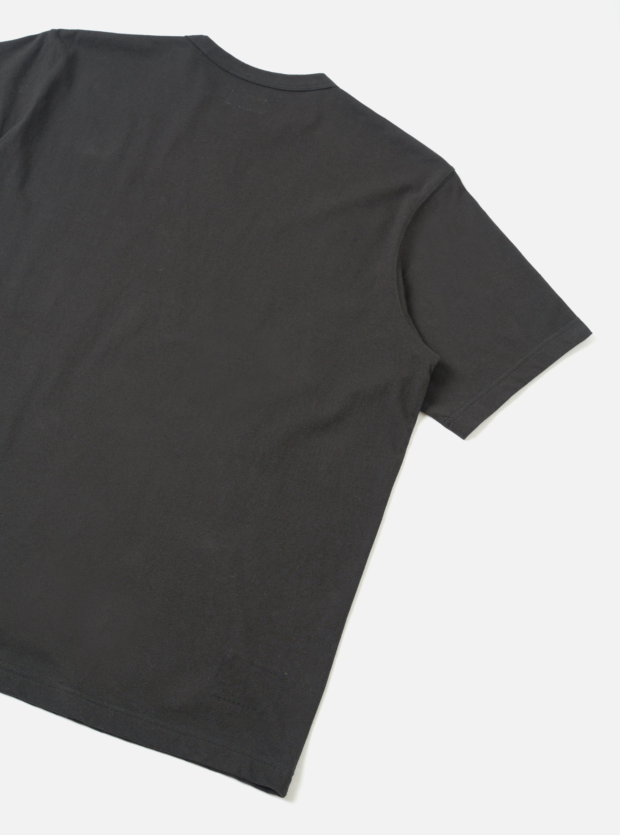 Back view of a black single jersey pocket tee, laid flat on a light grey surface. The shot is cropped to show the upper back, one short sleeve with a plain hem, and a care label visible at the nape of the neck. The fabric is smooth with a fine jersey knit texture.