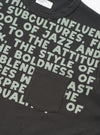 Close-up flat lay of a black cotton jersey pocket tee on a white studio surface. An all-over sage green bold text print covers the front. A single patch chest pocket sits at lower left. The ribbed crew neck and a Universal Works woven label are visible at the top.