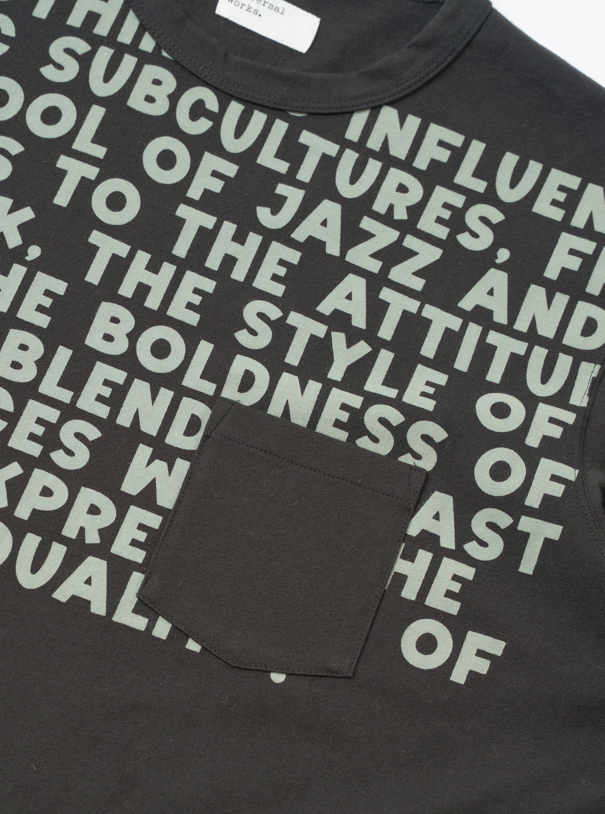 Close-up flat lay of a black cotton jersey pocket tee on a white studio surface. An all-over sage green bold text print covers the front. A single patch chest pocket sits at lower left. The ribbed crew neck and a Universal Works woven label are visible at the top.