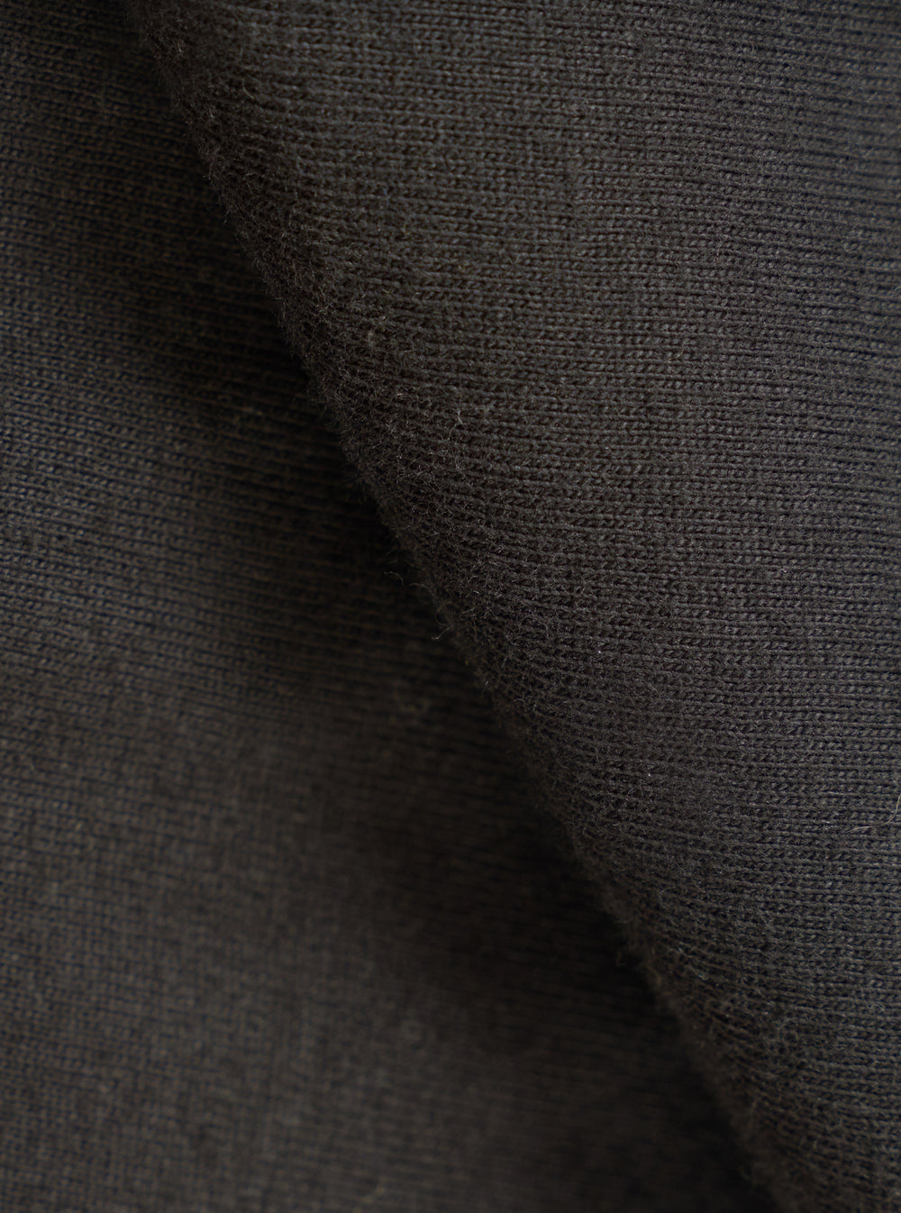 Close-up of the black single jersey knit fabric, showing a fine, even knit texture with a soft, slightly brushed surface. The fabric is folded to reveal depth and drape. No garment details or background are visible.