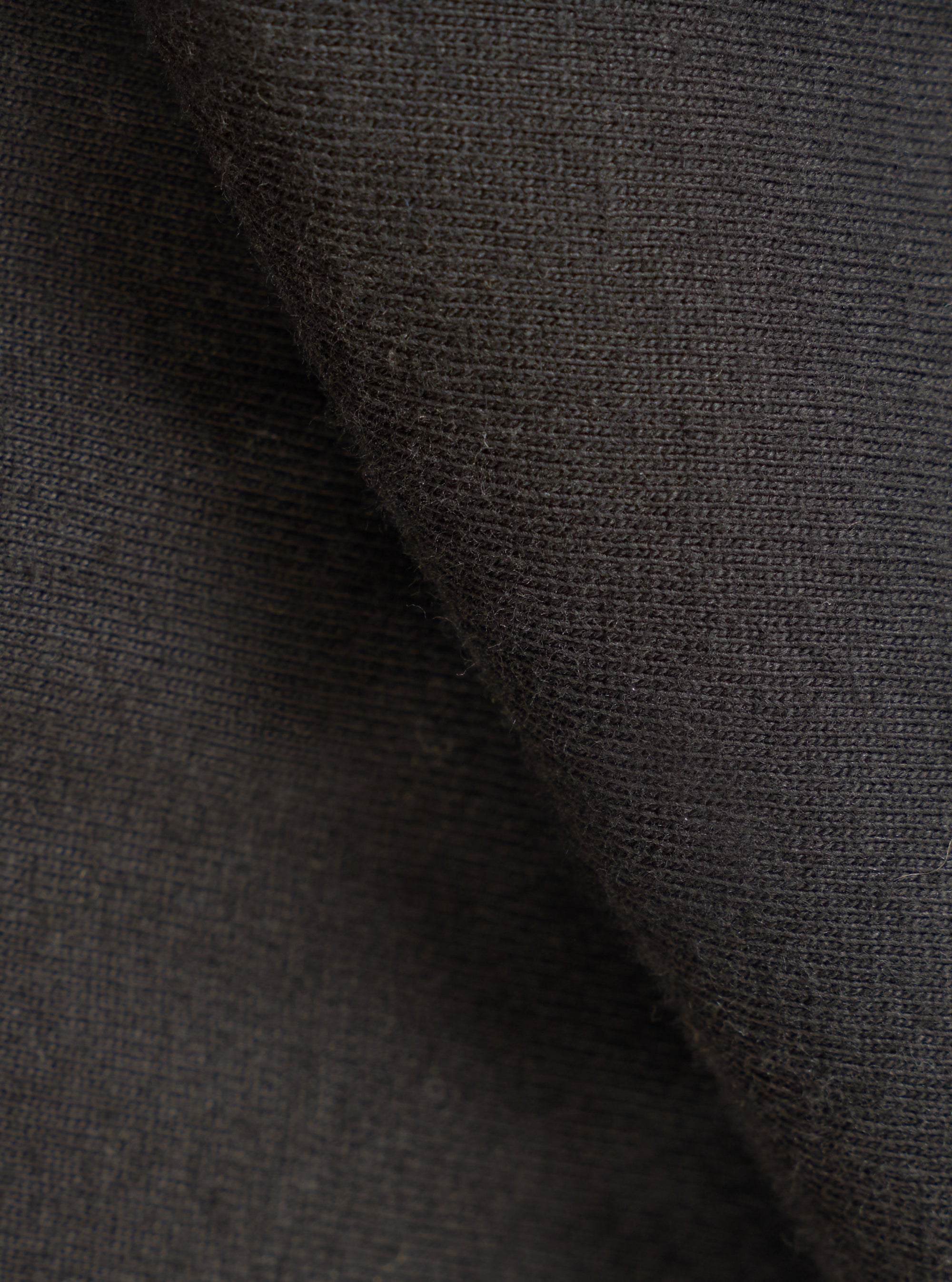 Close-up of the black single jersey knit fabric, showing a fine, even knit texture with a soft, slightly brushed surface. The fabric is folded to reveal depth and drape. No garment details or background are visible.