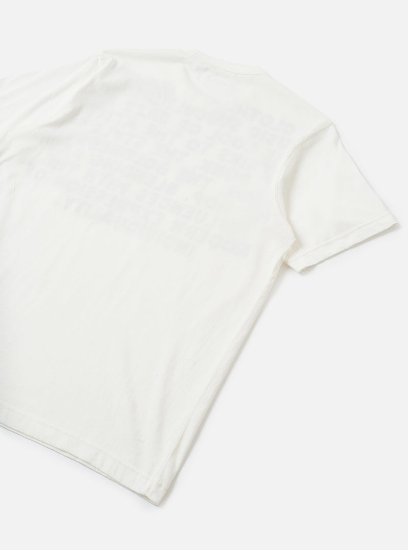 Flat lay of an ecru single jersey pocket tee on a white surface, shot from a diagonal angle showing the back and one short sleeve. The plain jersey fabric has a soft, lightweight texture with natural drape. A ribbed short sleeve cuff is visible on the right side.