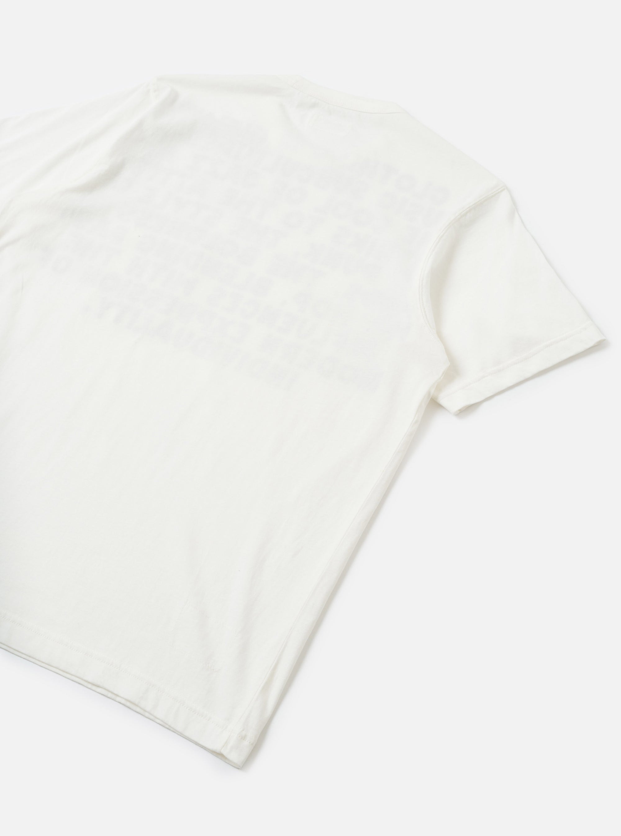 Flat lay of an ecru single jersey pocket tee on a white surface, shot from a diagonal angle showing the back and one short sleeve. The plain jersey fabric has a soft, lightweight texture with natural drape. A ribbed short sleeve cuff is visible on the right side.