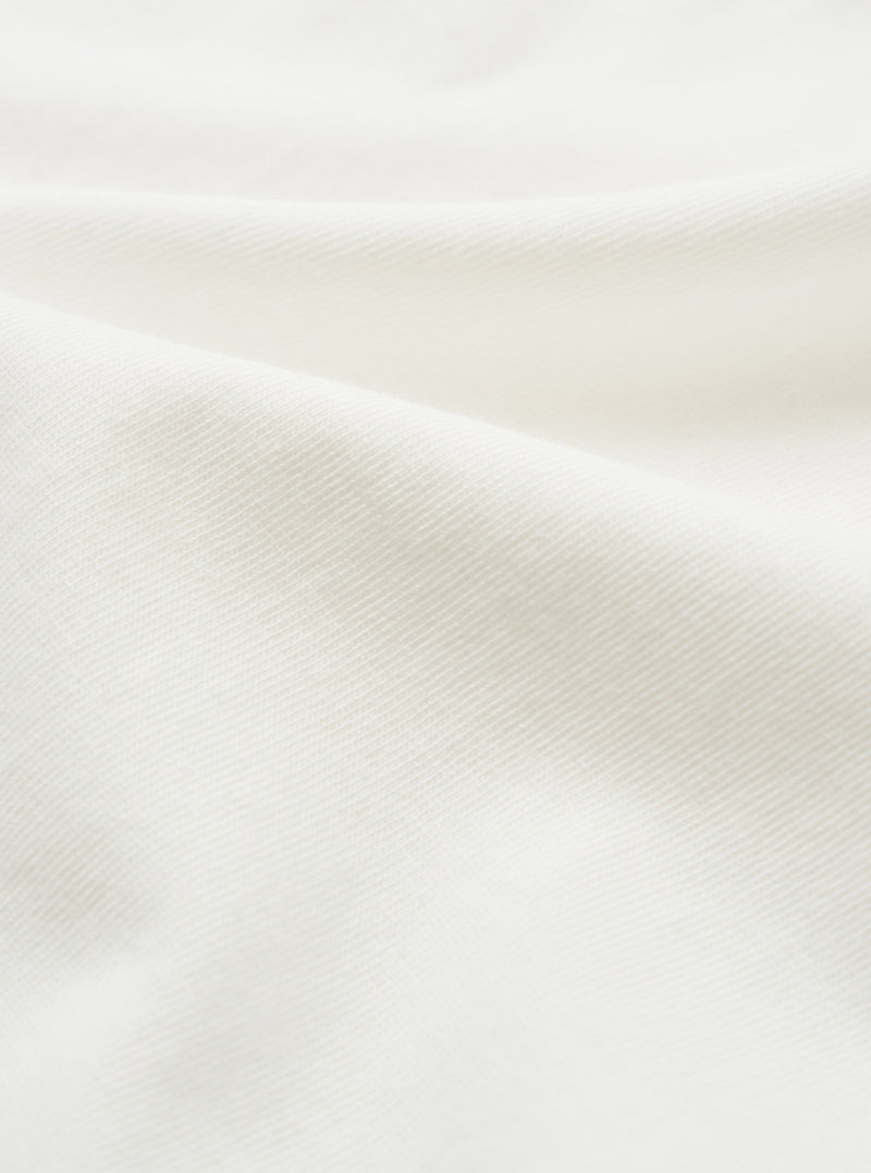 Close-up of the ecru single jersey fabric used in the Jazz Core Pocket Tee. The fine, smooth knit texture is visible across soft draped folds, showing a uniform diagonal weave structure in an off-white tone.