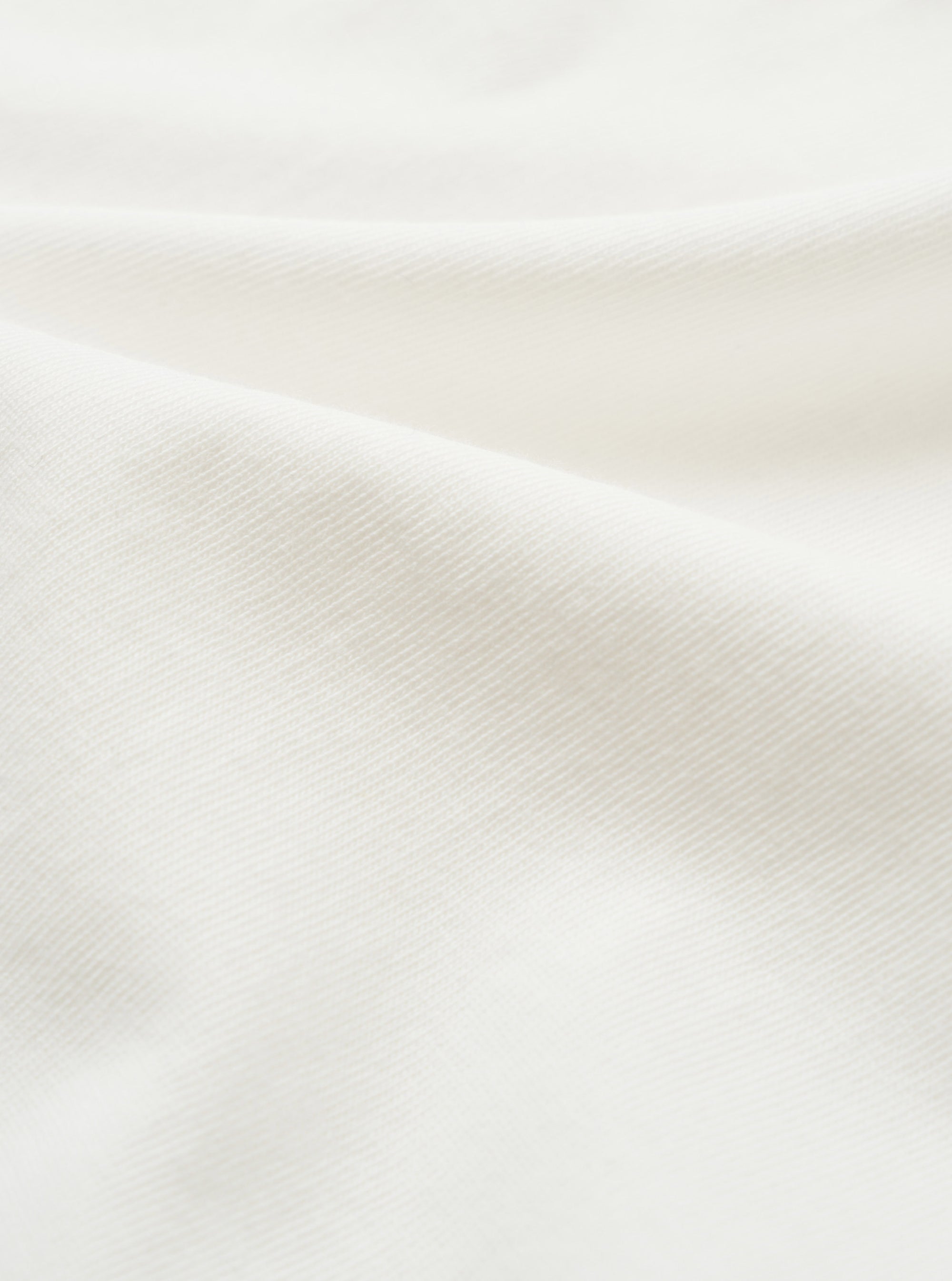 Close-up of the ecru single jersey fabric used in the Jazz Core Pocket Tee. The fine, smooth knit texture is visible across soft draped folds, showing a uniform diagonal weave structure in an off-white tone.