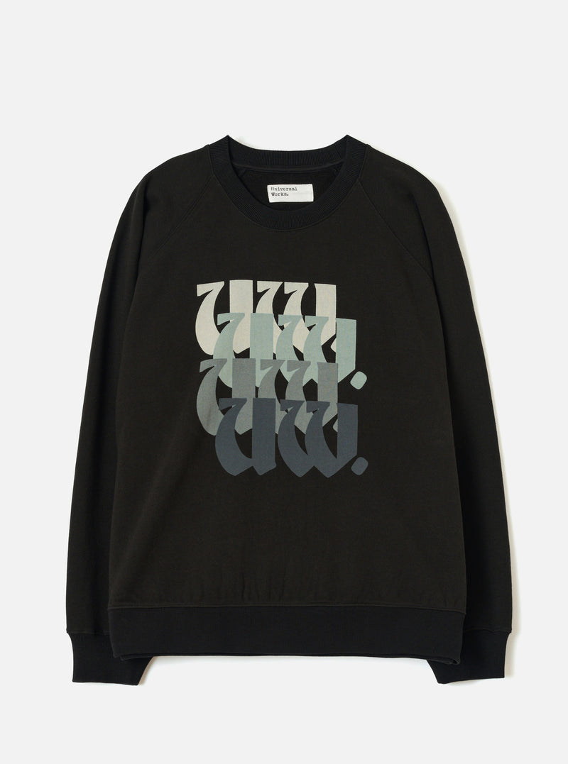 Black raglan sweatshirt laid flat on a light grey studio surface. Ribbed crew neck with a Universal Works woven label at the back neck. Ribbed cuffs and hem. Chest features a repeated "UW." graphic in blackletter style, fading from pale sage to dark charcoal across four stacked rows.