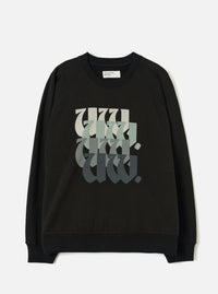 Black raglan sweatshirt laid flat on a light grey studio surface. Ribbed crew neck with a Universal Works woven label at the back neck. Ribbed cuffs and hem. Chest features a repeated "UW." graphic in blackletter style, fading from pale sage to dark charcoal across four stacked rows.