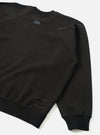 Black raglan sweatshirt laid flat on a white surface, shot from the back at an angle. Smooth brush back fabric with ribbed crew neck collar, ribbed hem and cuff. A small dark grey UW logo print sits below the back neckline. A white topstitched rectangular patch detail appears at the lower left hem.