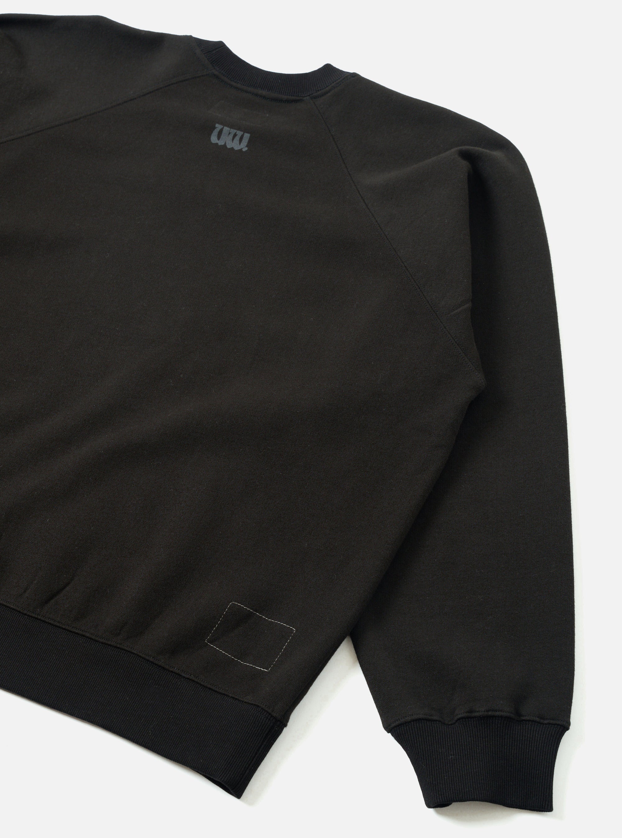 Black raglan sweatshirt laid flat on a white surface, shot from the back at an angle. Smooth brush back fabric with ribbed crew neck collar, ribbed hem and cuff. A small dark grey UW logo print sits below the back neckline. A white topstitched rectangular patch detail appears at the lower left hem.