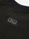 Close-up of a black raglan sweatshirt, focusing on the chest and ribbed crew neckline. A tonal dark grey gothic-style "UW!" logo print sits on the left chest. The raglan seam is visible across the upper chest. Flat lay on a white studio backdrop. Fleece-back fabric texture is visible.