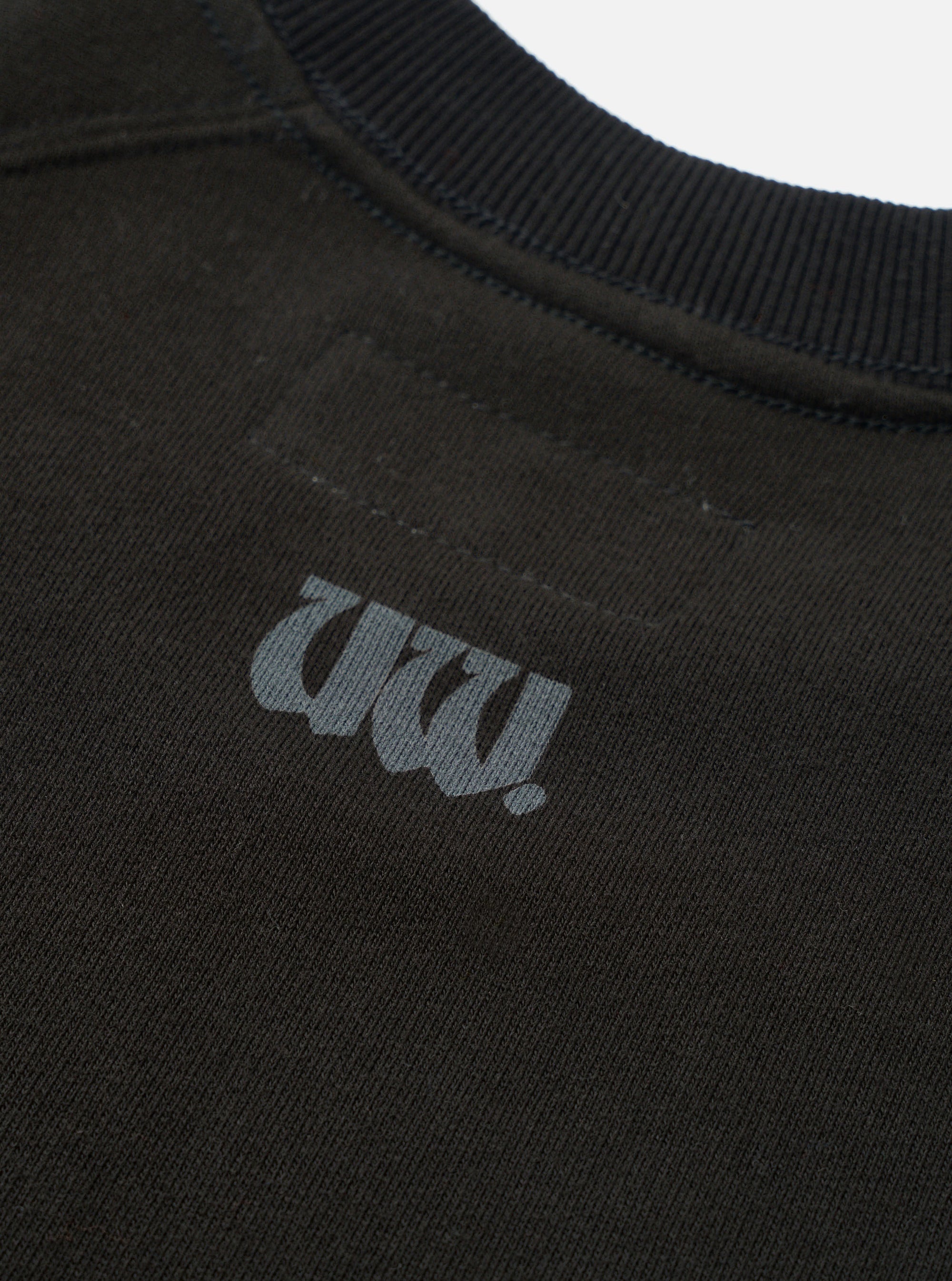 Close-up of a black raglan sweatshirt, focusing on the chest and ribbed crew neckline. A tonal dark grey gothic-style "UW!" logo print sits on the left chest. The raglan seam is visible across the upper chest. Flat lay on a white studio backdrop. Fleece-back fabric texture is visible.