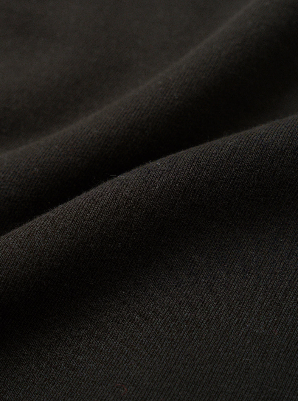 Close-up of black brushback fleece fabric, showing a soft, dense knit texture with a lightly napped surface. The fabric is folded and draped, revealing its weight and structure. No garment details or background are visible in this fabric detail shot.