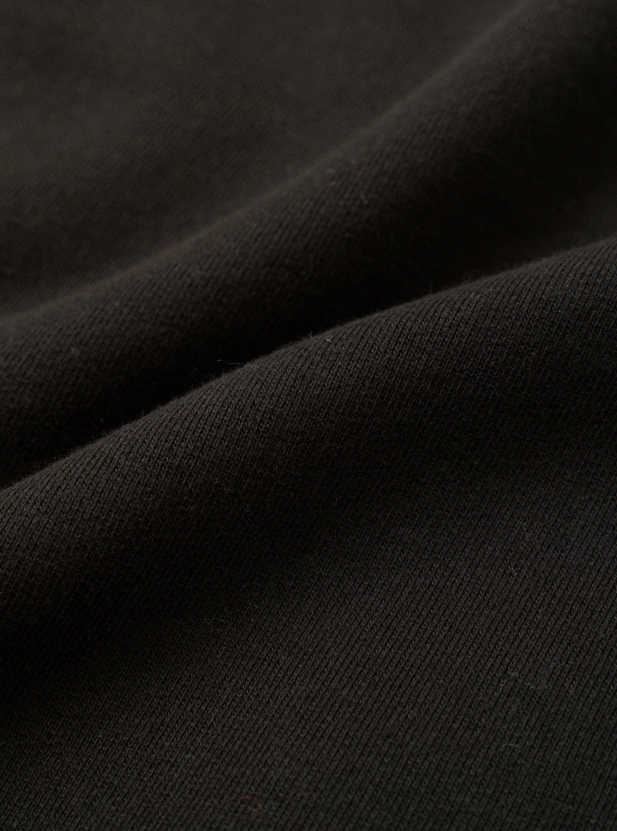 Close-up of black brushback fleece fabric, showing a soft, dense knit texture with a lightly napped surface. The fabric is folded and draped, revealing its weight and structure. No garment details or background are visible in this fabric detail shot.
