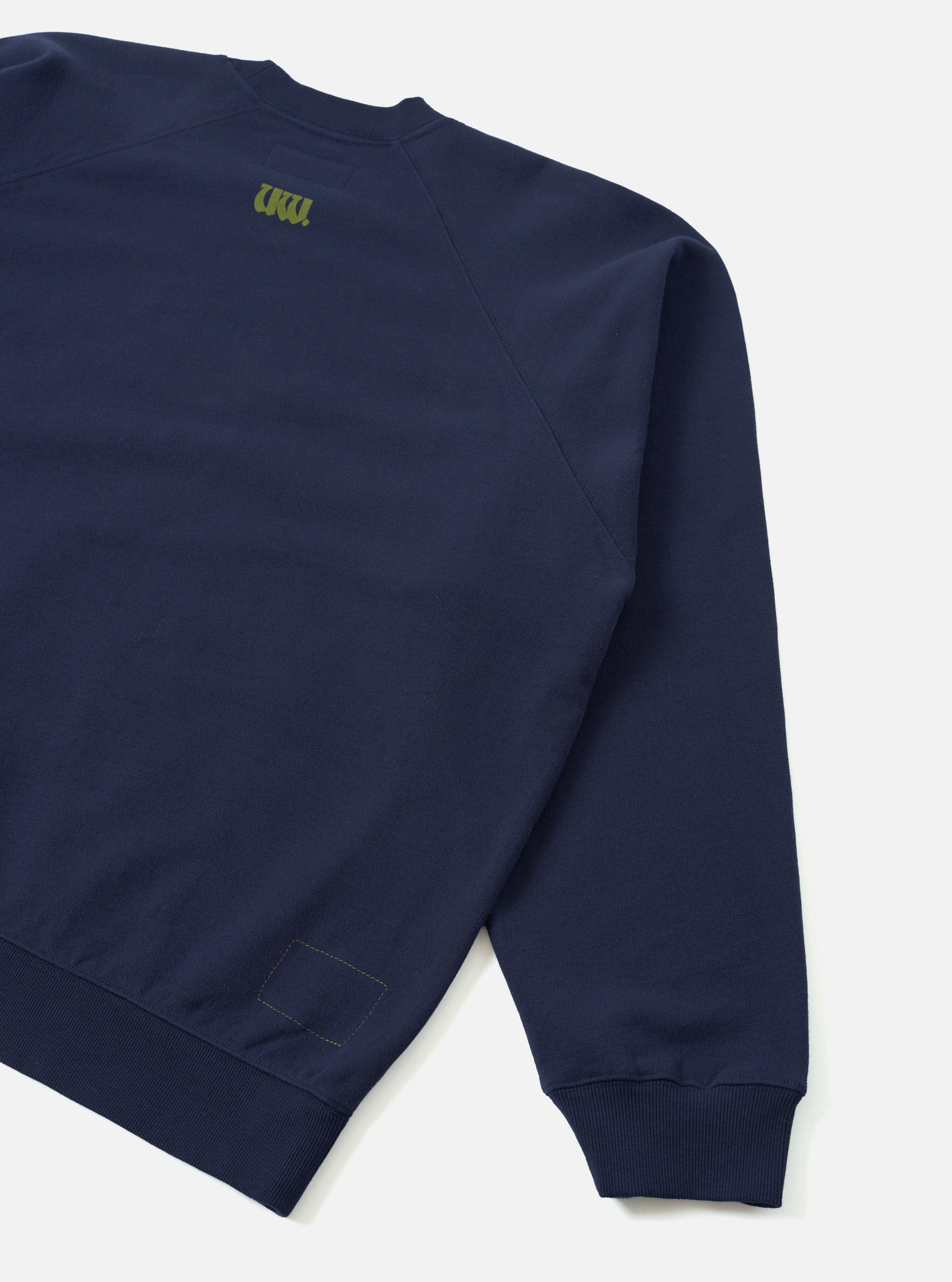 Navy brush back raglan sweatshirt, flat lay on a light grey seamless backdrop. Close-up of the back and left sleeve, showing the raglan seam, ribbed cuff and hem. A small olive green UW logo is printed at the centre back yoke. A stitched patch detail sits above the ribbed hem.