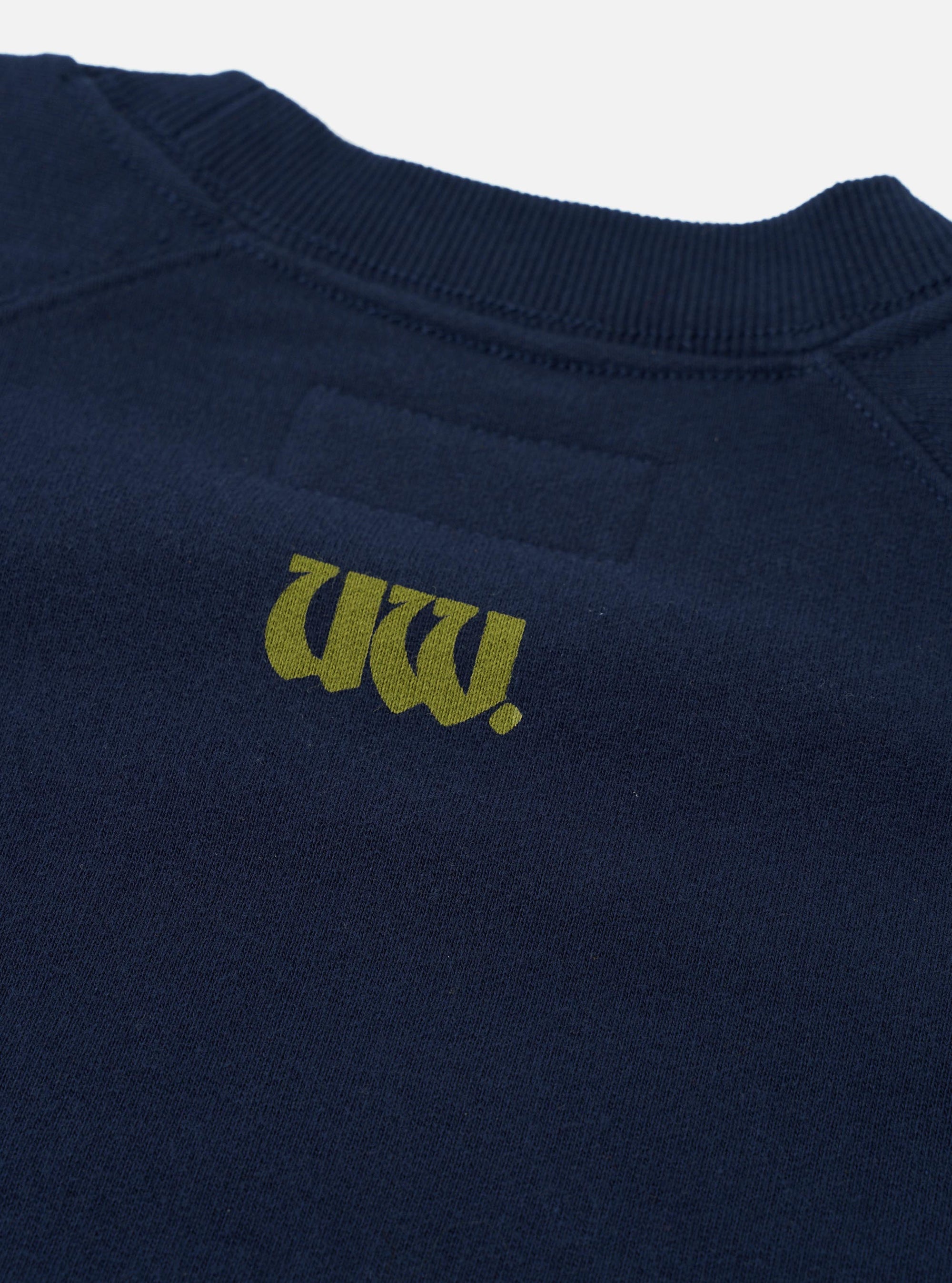 Close-up of a navy brush back raglan sweatshirt on a plain white background. A ribbed crew neck is visible at the top. The chest features a printed olive-yellow gothic-style "UW." logo. Raglan seam lines extend from the collar toward the body. The fleece fabric texture is visible throughout.