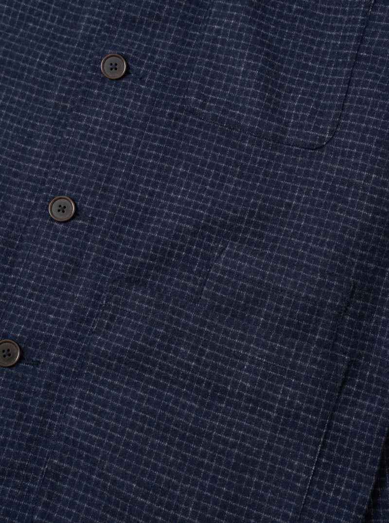Close-up of the navy check wool mix bakers overshirt, showing three dark horn-effect four-hole buttons along the front placket and a curved patch pocket at upper right. The fabric has a fine white grid check woven into a dark navy ground with a textured, slightly nubby handle. Plain white studio backdrop.