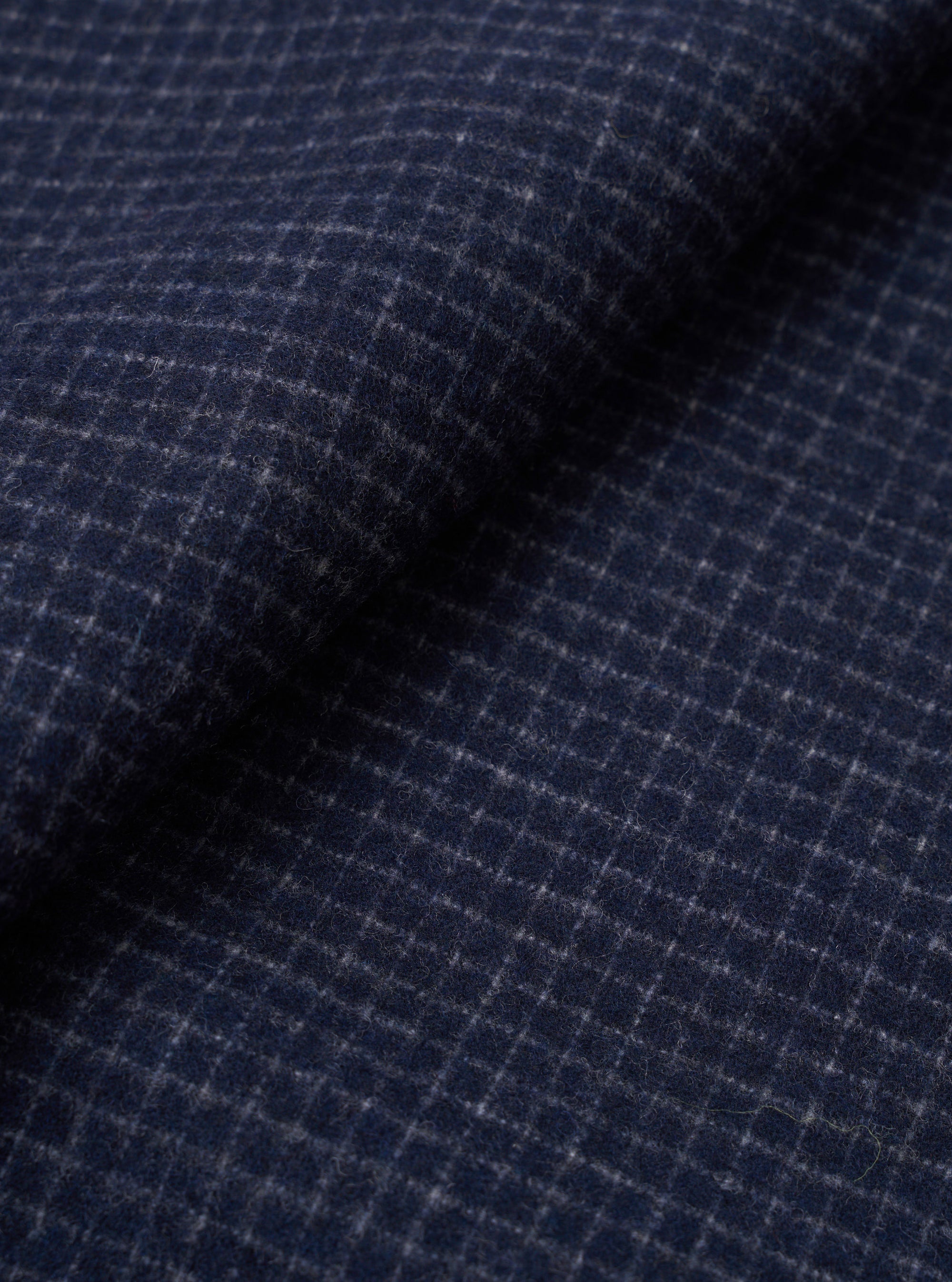 Close-up fabric shot of the navy check wool mix used in the Bobby Bakers Overshirt. A fine grey windowpane check runs across a navy ground, with a soft, lightly hairy wool texture visible across the draped and folded cloth.