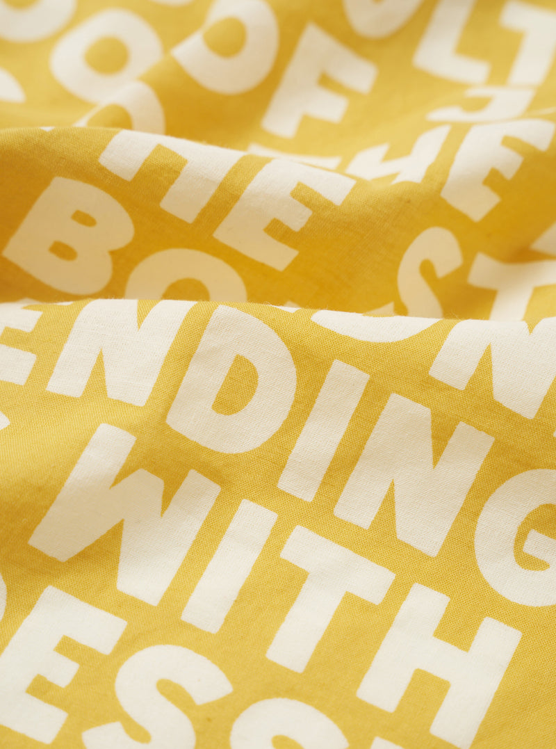 A close-up swatch image of the Mustard Jazz/Beatniks Print Neckerchief on a plain background, highlighting the mustard yellow cotton fabric and detailed jazz graphic text pattern.