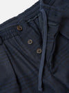 Close-up of the waistband area of the navy wool mix Oxford pant. A ribbed elasticated waistband sits above a button fly with three brown four-hole buttons. A navy drawcord threads through the waistband with a visible eyelet. The fabric shows a textured Oxford weave with a subtle dark check pattern. Plain white studio backdrop.