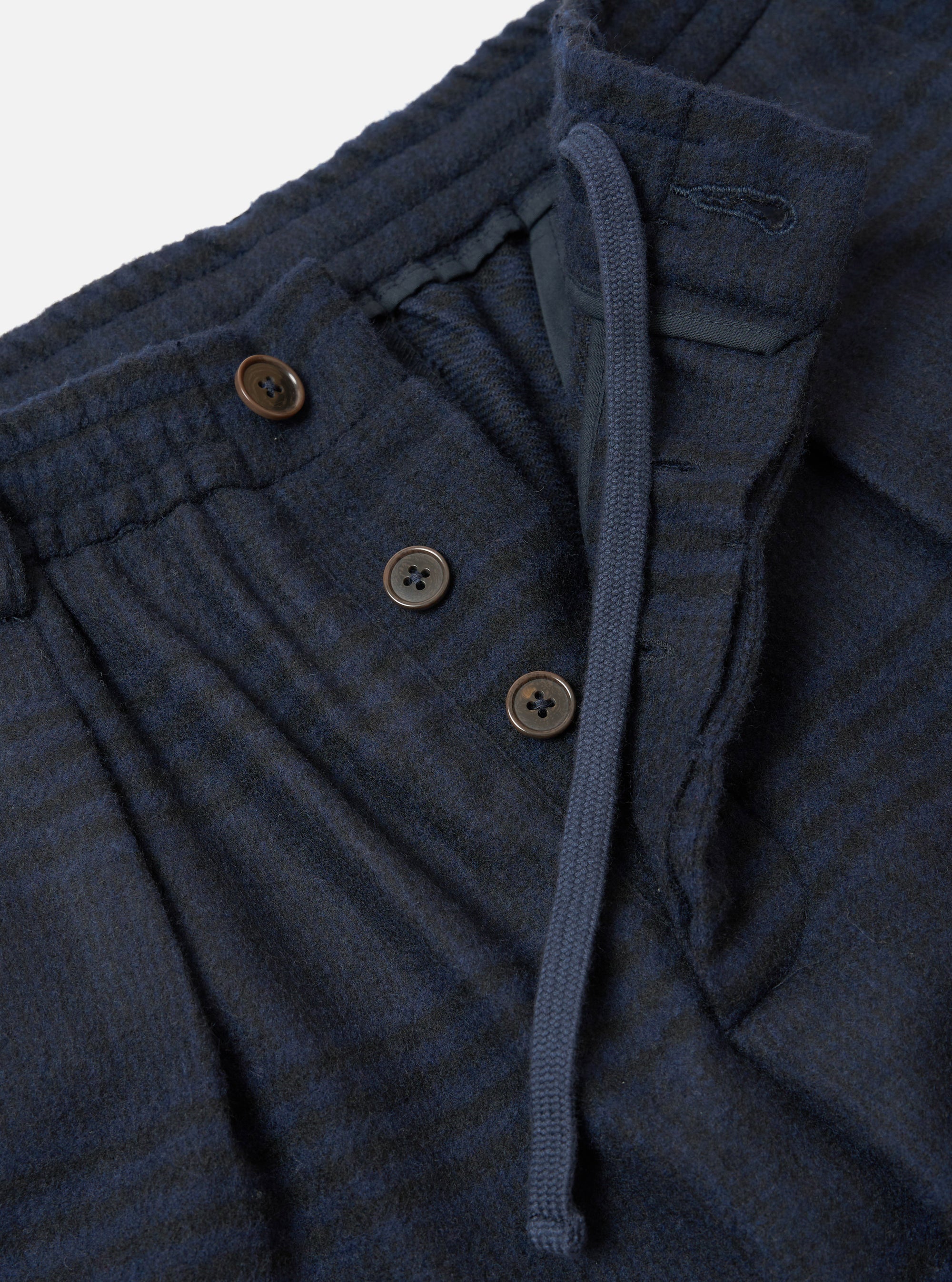 Close-up of the waistband area of the navy wool mix Oxford pant. A ribbed elasticated waistband sits above a button fly with three brown four-hole buttons. A navy drawcord threads through the waistband with a visible eyelet. The fabric shows a textured Oxford weave with a subtle dark check pattern. Plain white studio backdrop.