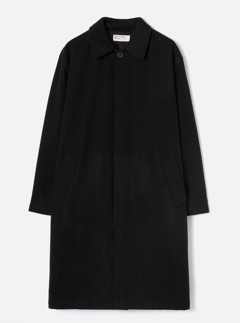 Black wool-look mac coat laid flat on a light grey studio backdrop. Features a point collar, concealed fly-front fastening with a single visible dark button at the neck, two patch pockets at the hip, and long plain sleeves. A Universal Works woven label is visible at the collar.