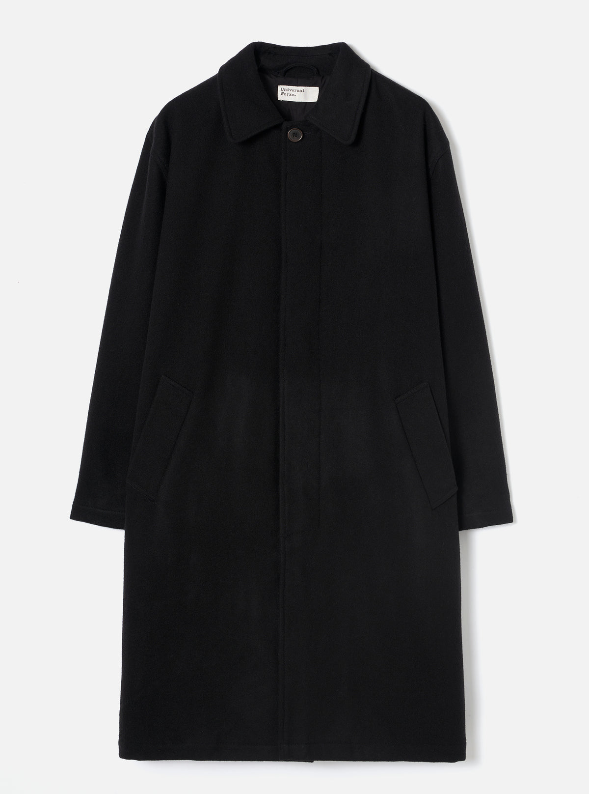 Black wool-look mac coat laid flat on a light grey studio backdrop. Features a point collar, concealed fly-front fastening with a single visible dark button at the neck, two patch pockets at the hip, and long plain sleeves. A Universal Works woven label is visible at the collar.