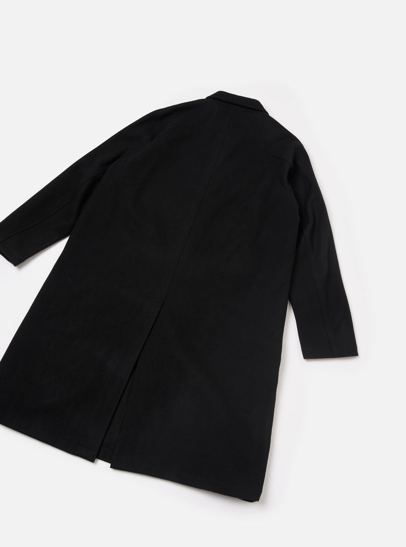 Black Mowbray Mac Coat laid flat on a light grey surface, shot from above at an angle showing the back. The smooth, matte fabric has a clean, unbroken back panel with a centre vent at the hem. A notched collar is visible at the top. Long sleeves with plain cuffs extend to the sides.