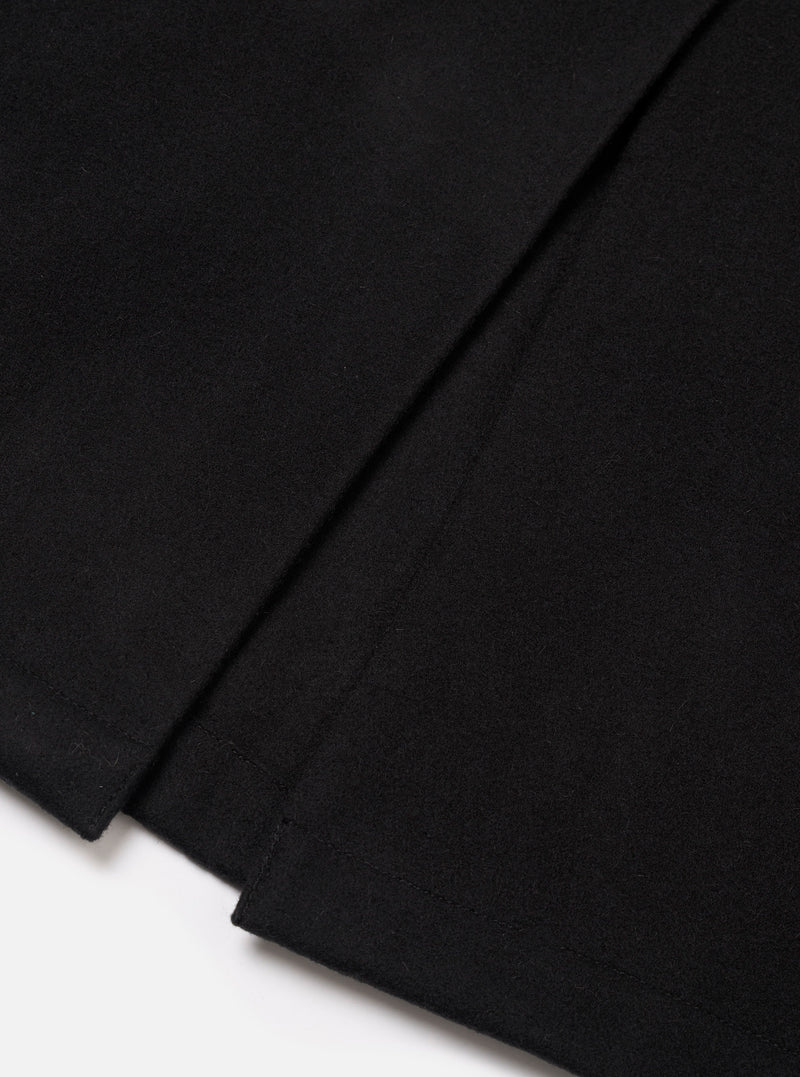 Close-up flat lay of the black Mowbray Mac Coat on a white surface, showing the lower hem and back vent detail. The black wool-blend fabric has a dense, lightly felted texture. The vent is stitched at the base with a clean, structured finish.