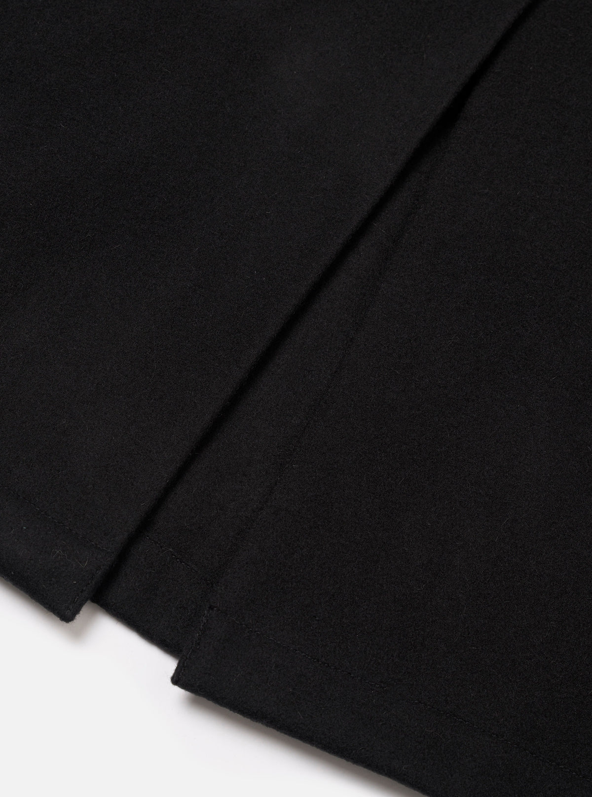 Close-up flat lay of the black Mowbray Mac Coat on a white surface, showing the lower hem and back vent detail. The black wool-blend fabric has a dense, lightly felted texture. The vent is stitched at the base with a clean, structured finish.