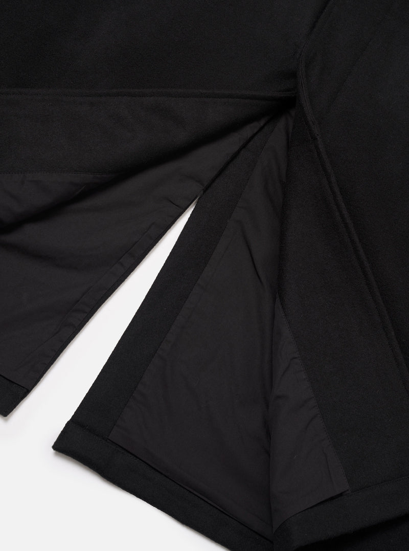 Close-up flat lay of the black Mowbray Mac Coat on a white surface, showing the open back vent detail. The matte wool-blend outer fabric contrasts with the smooth black satin lining visible inside the vent. Topstitched seams and a neatly finished hem are visible at the base of the coat.