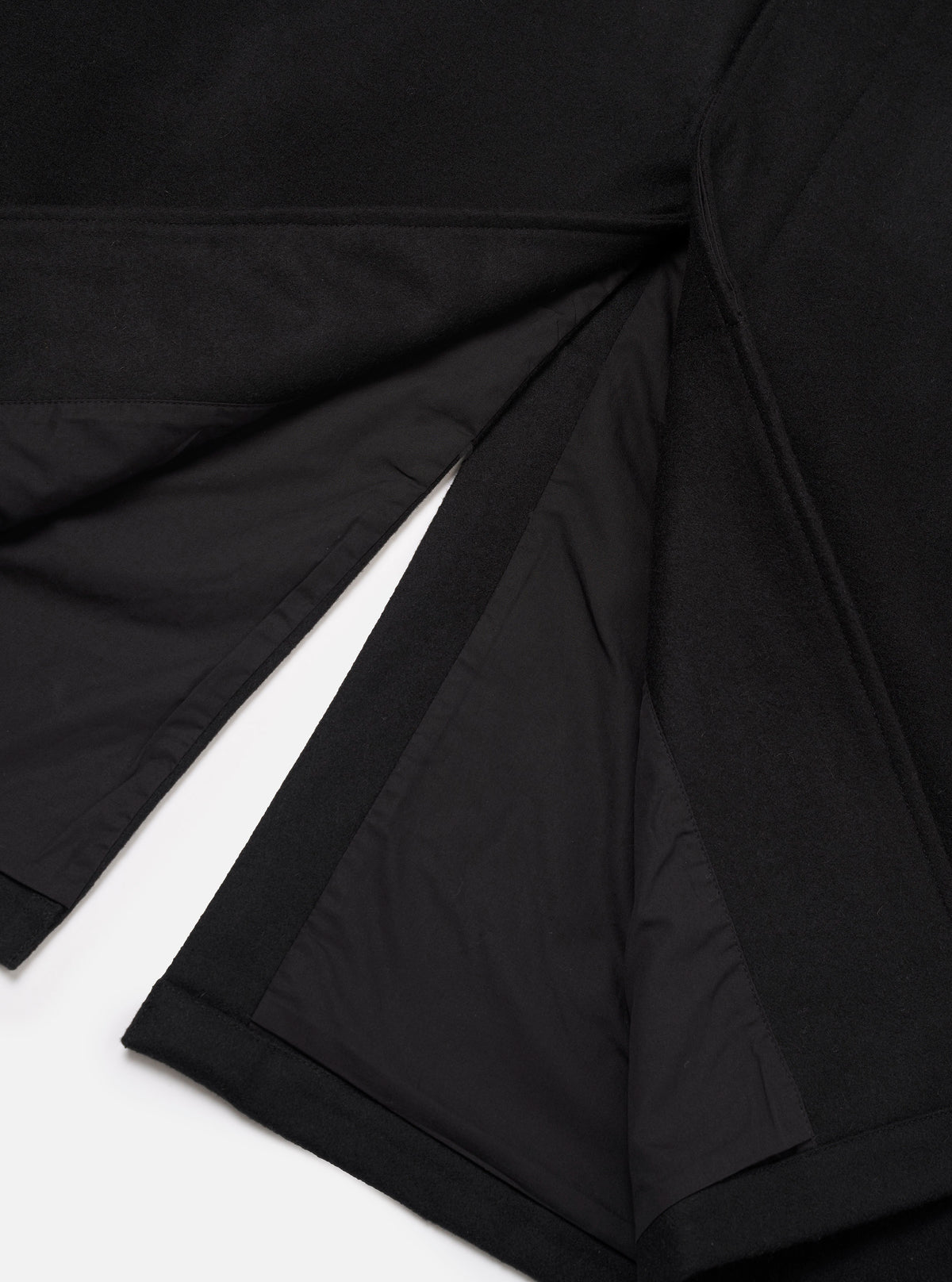 Close-up flat lay of the black Mowbray Mac Coat on a white surface, showing the open back vent detail. The matte wool-blend outer fabric contrasts with the smooth black satin lining visible inside the vent. Topstitched seams and a neatly finished hem are visible at the base of the coat.