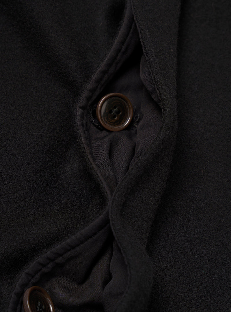 Close-up detail of the black Mowbray Mac Coat's button placket, showing two four-hole dark brown buttons against a soft, lightly textured wool-blend fabric. The front opening is partially open, revealing a dark woven lining with topstitched facing edges visible along the placket.
