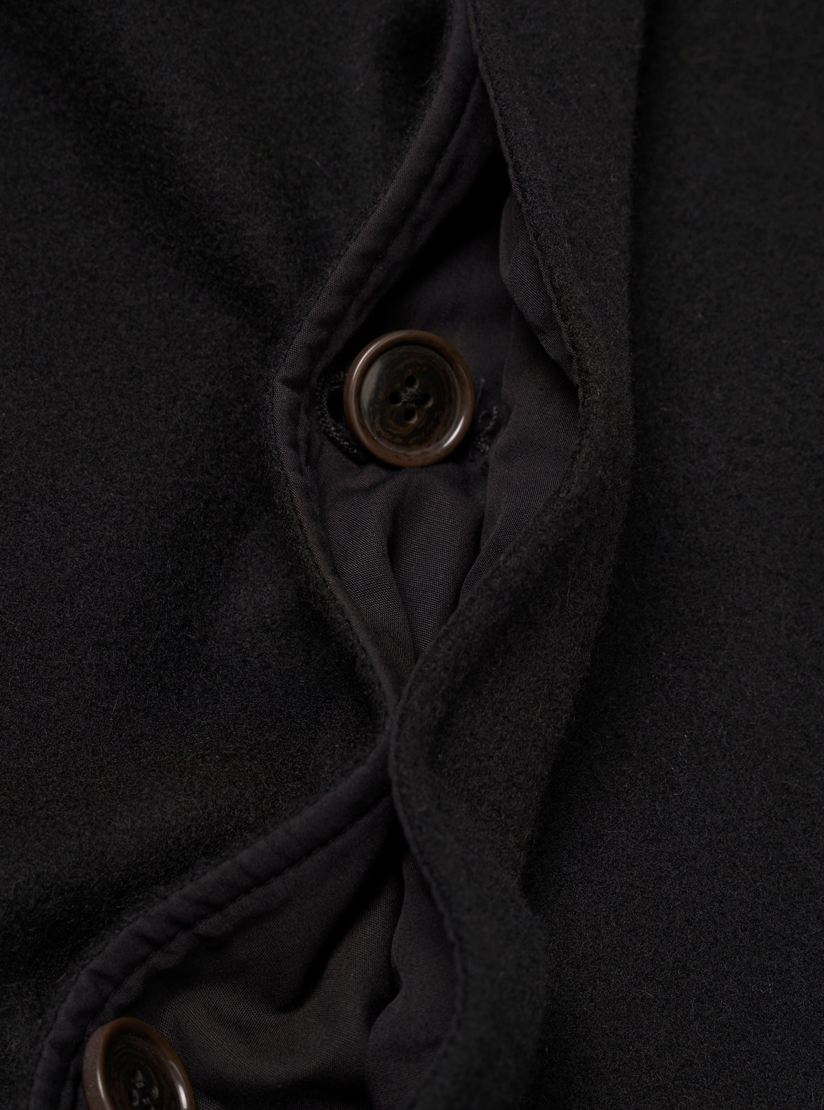 Close-up detail of the black Mowbray Mac Coat's button placket, showing two four-hole dark brown buttons against a soft, lightly textured wool-blend fabric. The front opening is partially open, revealing a dark woven lining with topstitched facing edges visible along the placket.