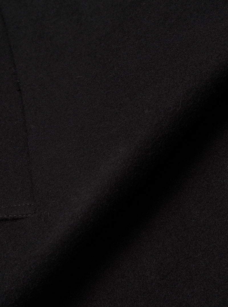 Close-up detail shot of the black Mowbray Mac Coat fabric. The tightly woven wool-blend cloth has a soft, lightly felted texture with a matte finish. A curved seam line is visible along the left edge, with fine topstitching. No background is visible.