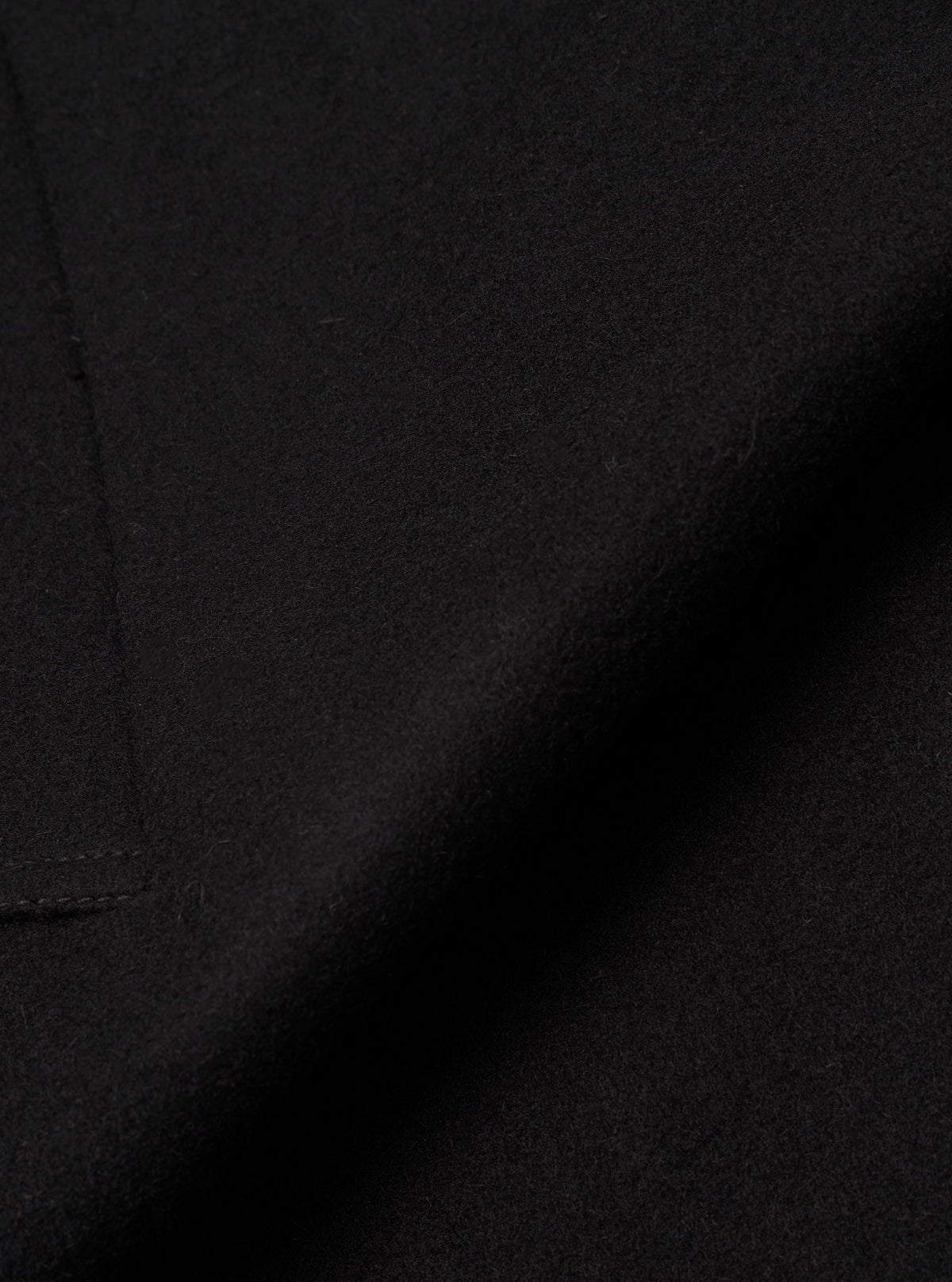 Close-up detail shot of the black Mowbray Mac Coat fabric. The tightly woven wool-blend cloth has a soft, lightly felted texture with a matte finish. A curved seam line is visible along the left edge, with fine topstitching. No background is visible.