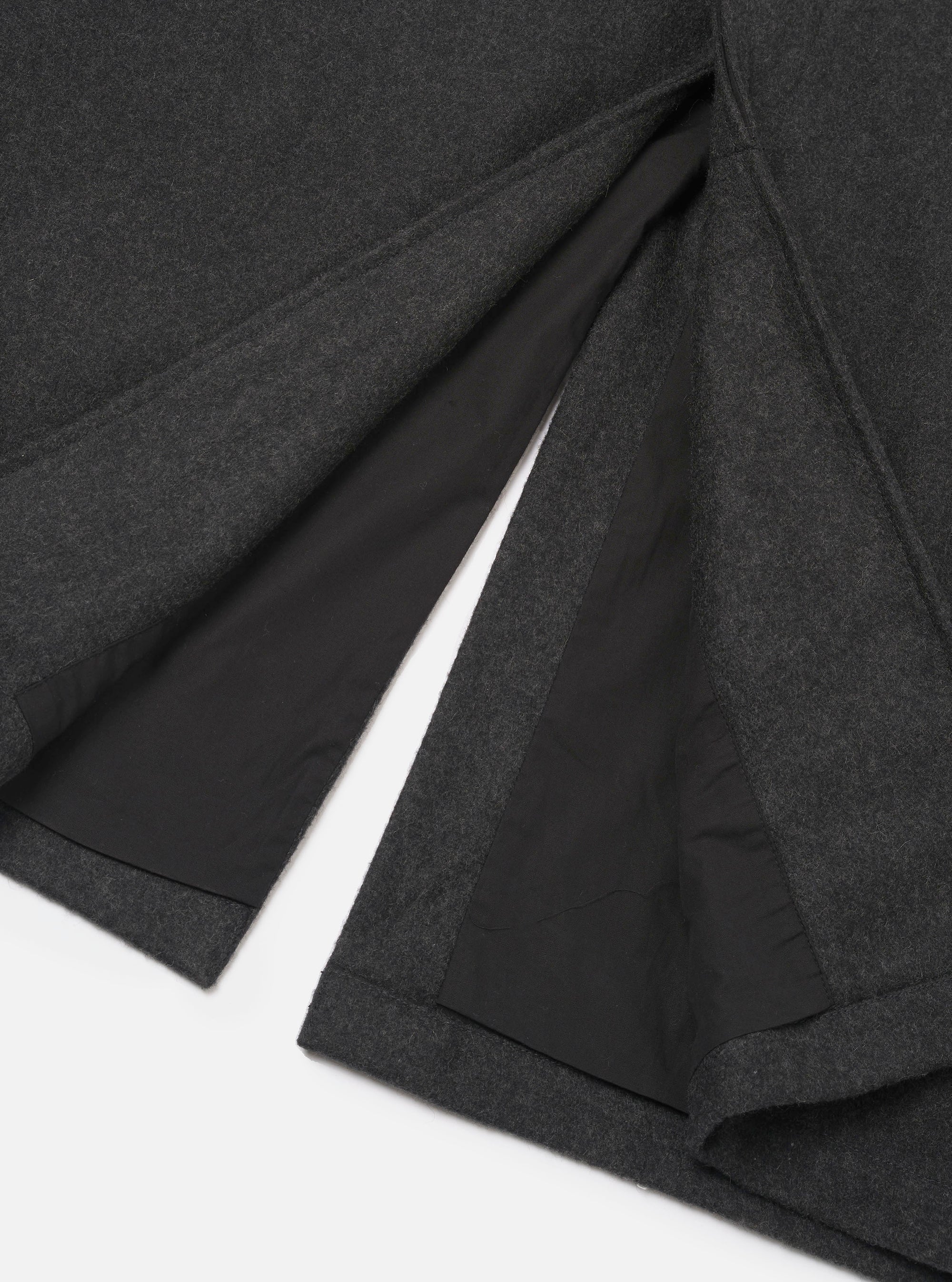 Close-up flat lay of the grey marl Mowbray Mac Coat hem and vent detail on a white surface. The back vent is open, revealing a black lining. The outer fabric is a medium-weight felted wool in dark charcoal marl. Topstitched edges finish the hem and vent cleanly.