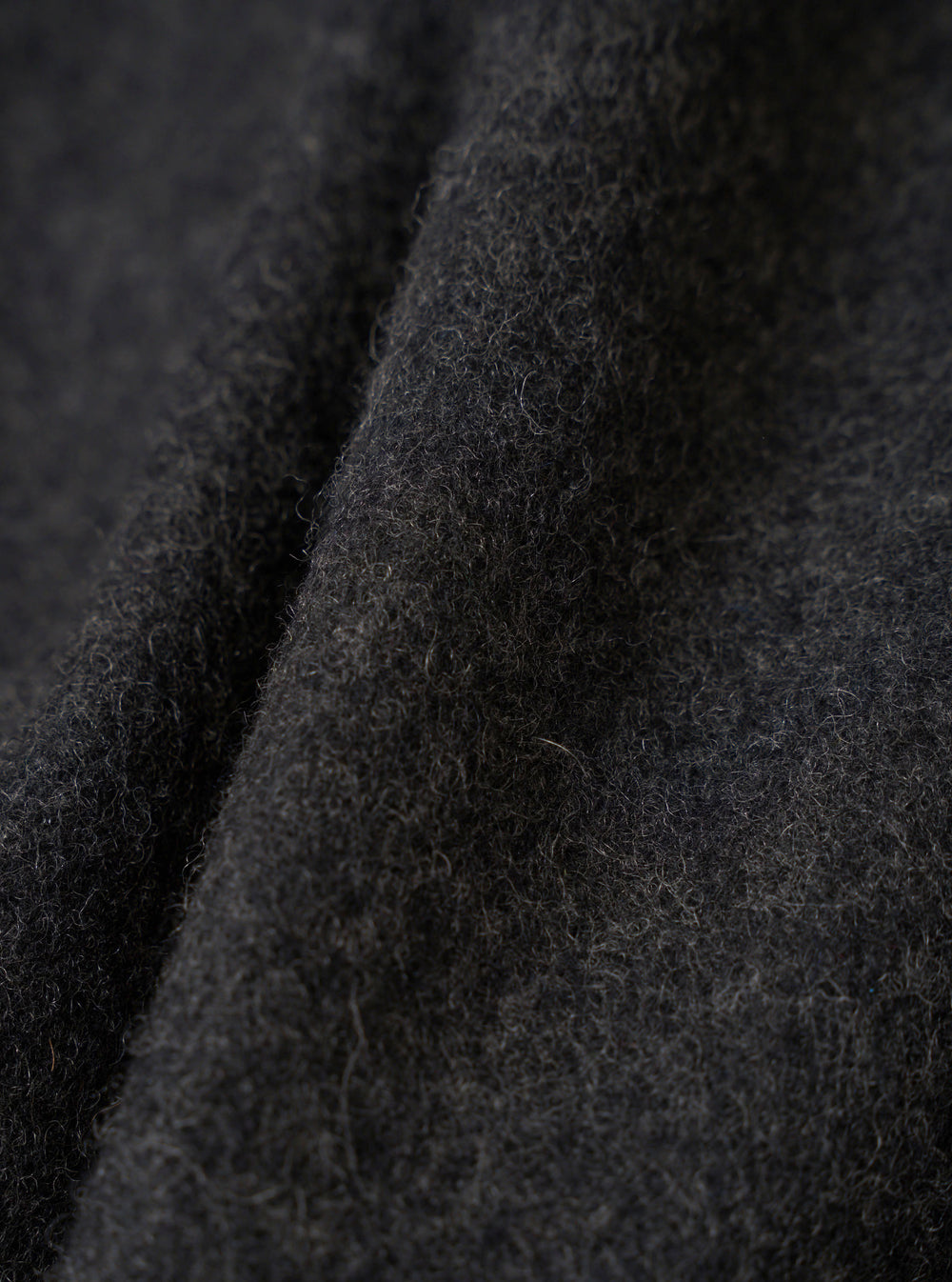 Close-up detail shot of the grey marl fabric on the Mowbray Mac Coat. The brushed wool textile shows a dense, fluffy surface with fine curled fibres and a dark charcoal tone with lighter grey flecks throughout. Soft folds reveal the fabric's weight and texture against a dark background.