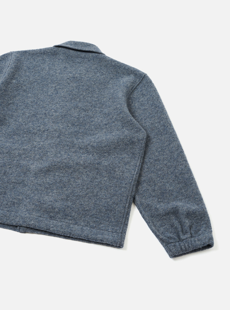 A back view of the Slate Blue Wool Fleece Rams Jacket displayed flat on a simple studio backdrop. The unadorned rear cut emphasizes the soft fleece finish, straight hem, and relaxed boxy construction.